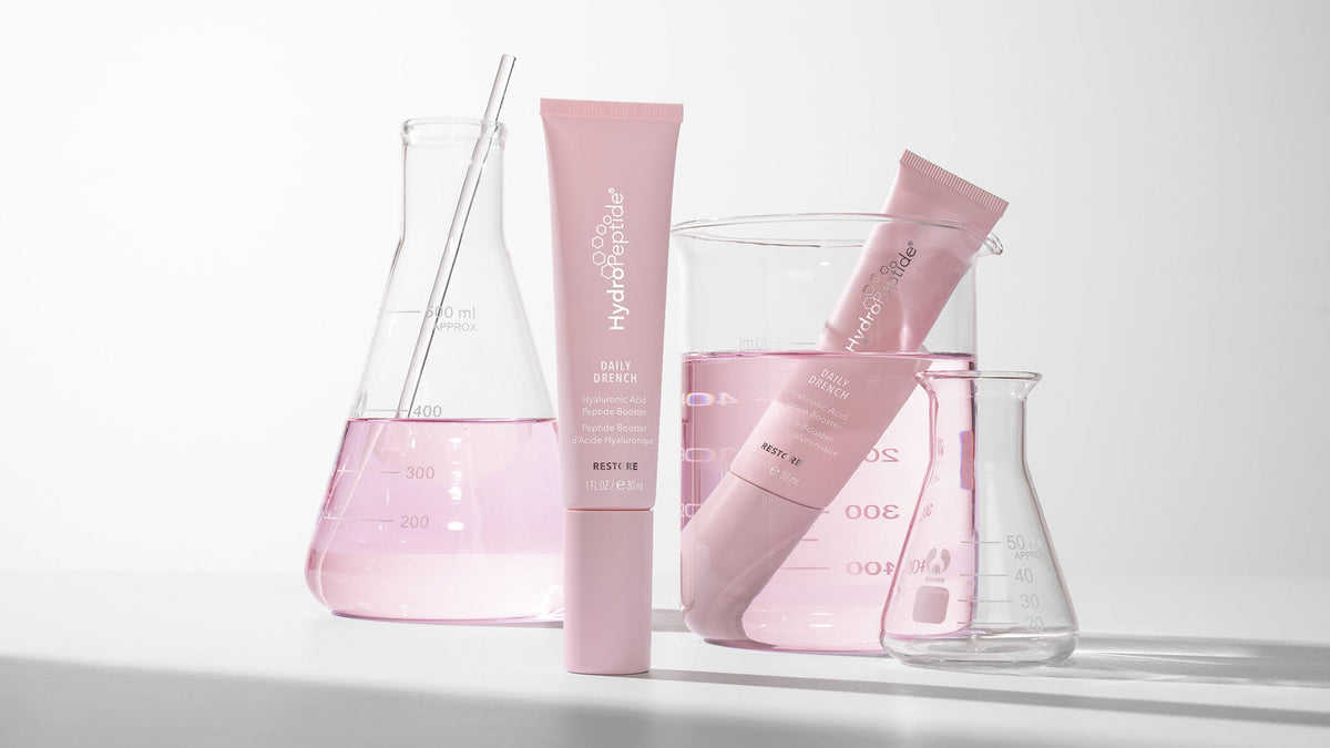 Two light pink tubes of Daily Drench against clear glass beakers with light pink liquid