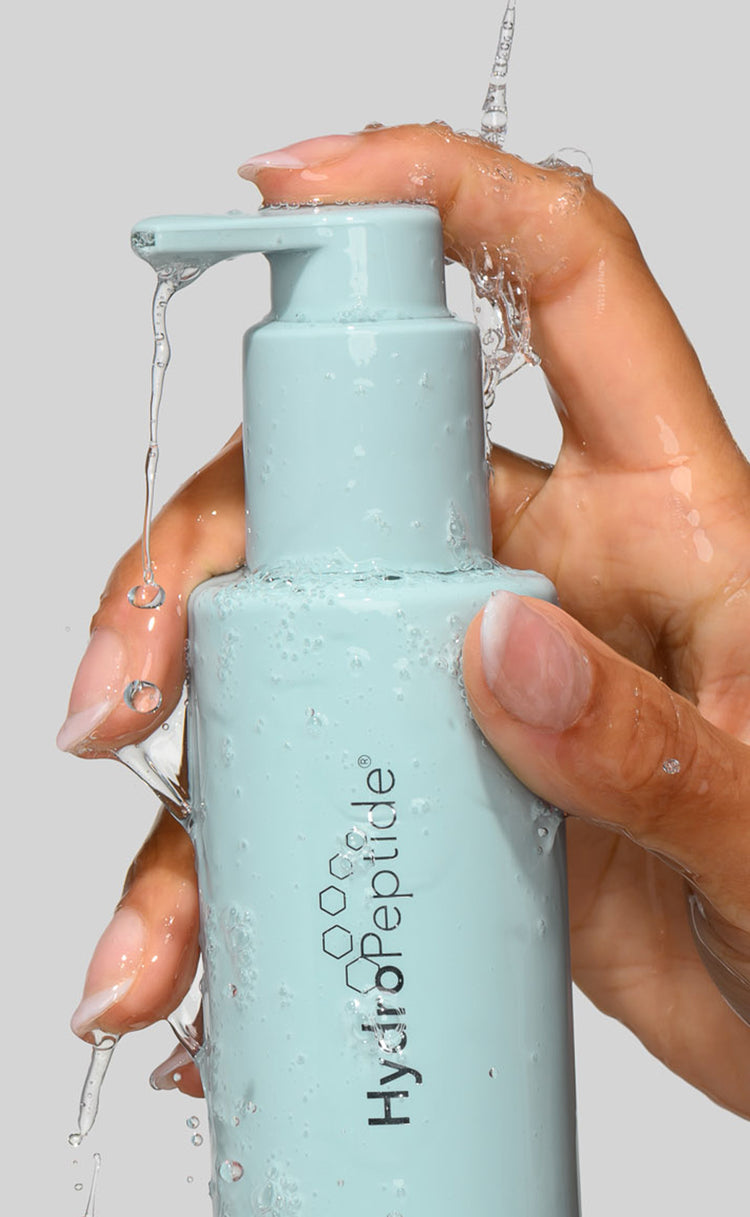 A hand holding a light blue pump bottle labeled 'HydroPeptide' with water dripping from the pump and surrounding the bottle, against a plain light gray background.